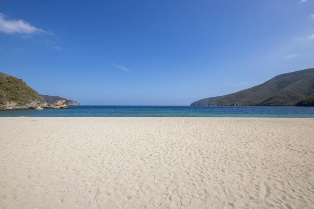 Serene beach scene with clear blue waters and lush green hills under a bright sky, perfect for a summer getaway., # **Tayrona: guía completa para visitar el Parque Nacional Natural Tayrona**El **Parque Nacional Natural Tayrona** es uno de los destinos naturales más representativos del Caribe colombiano. En este territorio encontramos playas rodeadas de montañas, senderos entre bosques tropicales, formaciones rocosas, arrecifes coralinos y espacios de enorme valor espiritual para los pueblos indígenas de la Sierra Nevada de Santa Marta.Visitar el Tayrona no consiste únicamente en pasar un día frente al mar. La experiencia combina **senderismo, contemplación de la naturaleza, contacto con ecosistemas marino-costeros, observación de fauna, cultura ancestral y turismo responsable**. Por esta razón, debemos planificar el recorrido con anticipación, elegir correctamente el acceso y conocer las restricciones establecidas para proteger el área.El parque forma parte de la **Línea Negra**, territorio ancestral de los pueblos Kogui, Wiwa, Arhuaco y Kankuamo. Dentro del área protegida existen sitios sagrados conectados cultural, espiritual y ecológicamente con la Sierra Nevada de Santa Marta, por lo que nuestra visita debe desarrollarse con respeto por las comunidades, los senderos autorizados y las normas de conservación.## **Dónde queda el Parque Tayrona**El **Parque Tayrona está ubicado en Santa Marta**, departamento del Magdalena, sobre la costa norte de Colombia y en las estribaciones de la Sierra Nevada de Santa Marta. Su posición geográfica permite que, en un mismo paisaje, confluyan montañas, bosques, playas, bahías, manglares, arrecifes y zonas marinas.El acceso más utilizado es **El Zaino**, situado aproximadamente a 32 kilómetros de Santa Marta por la Troncal del Caribe en dirección a Riohacha. Desde el centro de la ciudad, el recorrido terrestre suele tomar alrededor de 45 minutos, aunque el tiempo puede aumentar según el tráfico, la temporada turística y las condiciones de la carretera. Desde El Zaino podemos dirigirnos hacia Cañaveral, Arrecifes y Cabo San Juan del Guía.También encontramos entradas por **Calabazo, Palangana y Bahía Concha**, cada una conectada con sectores diferentes. Por ello, antes de desplazarnos debemos definir qué playa, sendero o experiencia deseamos conocer.## **Cómo llegar al Tayrona desde Santa Marta**### **Entrada por El Zaino**El Zaino es la entrada principal y la alternativa más recomendable cuando deseamos realizar la ruta clásica hacia **Cañaveral, Arrecifes y Cabo San Juan**. Después de ingresar, podemos utilizar el transporte interno disponible hasta el sector de Cañaveral y continuar caminando por los senderos habilitados.Esta ruta exige tiempo, buena condición física y calzado apropiado. Aunque gran parte del recorrido se realiza bajo vegetación, existen tramos expuestos al calor, zonas húmedas, pasarelas, pendientes y superficies irregulares.### **Entrada por Calabazo**Calabazo se encuentra en el kilómetro 20 de la Troncal del Caribe. Desde este acceso podemos caminar hacia **Playa Brava, Boca del Saco y Cabo San Juan del Guía**. Es una alternativa asociada a recorridos más largos y exigentes, por lo que debemos ingresar temprano, llevar equipaje ligero y respetar los horarios de salida.### **Entrada por Palangana**El acceso por Palangana permite dirigirnos hacia **Gayraca, Neguanje, Playa del Muerto, Playa Cristal y Cinto**. Algunas de estas playas requieren combinar el desplazamiento terrestre con transporte en lancha y funcionan principalmente bajo modalidad de pasadía.Esta entrada resulta adecuada cuando buscamos disfrutar del mar sin realizar la caminata extensa que caracteriza la ruta de El Zaino.### **Entrada por Bahía Concha**Bahía Concha es uno de los sectores más cercanos a la zona urbana de Santa Marta. Después de llegar al portón de entrada, debemos realizar una caminata de aproximadamente 20 minutos hasta la playa principal. Desde este sector también pueden operar recorridos marítimos hacia playas cercanas, según las condiciones autorizadas.### **Ingreso marítimo al Parque Tayrona**También podemos llegar por vía marítima desde sectores como **Santa Marta, El Rodadero o Taganga**, con rutas hacia Granate, Bahía Concha, Neguanje, Cinto y Cabo San Juan. Antes de contratar una embarcación debemos verificar que el operador esté autorizado, conocer el estado del mar y confirmar que el servicio incluya las condiciones de ingreso y el seguro correspondiente.## **Mejores playas del Parque Tayrona**### **Cabo San Juan del Guía****Cabo San Juan** es uno de los lugares más conocidos del Tayrona. Se distingue por sus dos playas separadas por una formación rocosa coronada por un kiosco. El paisaje reúne arena, palmeras, bosque tropical, grandes piedras y aguas caribeñas.La ruta habitual desde El Zaino atraviesa Cañaveral, Arrecifes y diferentes tramos de bosque costero. El sendero Arrecifes–Cabo San Juan permite realizar actividades de observación de flora y fauna, fotografía, interpretación del patrimonio y natación en las zonas autorizadas.Por su popularidad, Cabo San Juan puede recibir una alta cantidad de visitantes durante vacaciones, fines de semana y días festivos. Recomendamos iniciar el recorrido temprano para caminar con menor exposición al calor y disponer de suficiente tiempo para regresar antes del cierre.### **Arrecifes**Arrecifes es uno de los paisajes más impactantes del parque por la fuerza del mar, las formaciones rocosas y la vegetación que rodea la costa. En este sector encontramos servicios de alojamiento, camping y alimentación autorizados, además de conexiones con los senderos hacia Cabo San Juan.No debemos asumir que todas las playas del Tayrona son aptas para nadar. El ingreso al mar únicamente debe realizarse en las zonas habilitadas y cuando la señalización lo permita. Las playas con **bandera roja permanecen cerradas por seguridad**.### **La piscina natural del sendero Arrecifes–Cabo San Juan**En el recorrido hacia Cabo San Juan encontramos un sector protegido por formaciones naturales que disminuyen la fuerza del oleaje. Esta zona permite la práctica de natación libre y careteo cuando las condiciones del parque lo autorizan.Aunque el agua pueda parecer tranquila, siempre debemos atender la señalización, las advertencias del personal y las condiciones climáticas del día.### **Neguanje y Playa Cristal**Neguanje funciona como punto de conexión hacia diferentes playas del sector de Palangana. Desde allí podemos desplazarnos por vía marítima hacia **Playa Cristal**, conocida por sus aguas claras y por las posibilidades de observar vida marina mediante actividades autorizadas.El acceso depende de la capacidad diaria, las condiciones del mar y la operación de las embarcaciones. Conviene reservar con anticipación y contratar únicamente prestadores habilitados.### **Bahía Concha**Bahía Concha ofrece una experiencia diferente a la ruta de senderismo hacia Cabo San Juan. Su cercanía con Santa Marta y su acceso relativamente corto la convierten en una alternativa adecuada para quienes disponen de menos tiempo o prefieren reducir la exigencia física.En este sector están autorizadas actividades como natación libre y buceo con equipo autónomo o a pulmón, siempre dentro de las áreas permitidas y bajo las condiciones de seguridad establecidas.### **Playa Brava**Playa Brava se encuentra conectada con el acceso de Calabazo y requiere una caminata exigente. Es una opción apropiada cuando buscamos una experiencia de senderismo más intensa y un ambiente menos concurrido.Debemos verificar previamente la disponibilidad de alojamiento, las condiciones del sendero y los horarios de ingreso. No es recomendable iniciar esta ruta tarde, improvisar el recorrido ni caminar fuera de las zonas señalizadas.## **Qué hacer en el Parque Nacional Natural Tayrona**### **Realizar senderismo ecológico**El senderismo es una de las principales actividades del Tayrona. Podemos recorrer rutas costeras, bosques, miradores y zonas de transición entre ecosistemas terrestres y marinos.La **Ruta del Conocimiento entre Cañaveral y Arrecifes** atraviesa espacios de interpretación ambiental, escenarios para observar fauna y flora y diferentes miradores naturales. El recorrido debe realizarse exclusivamente por los caminos habilitados.### **Observar fauna y flora**El parque protege bosques secos, bosques húmedos, manglares, playas, fondos marinos, arrecifes y otros ecosistemas que sostienen una elevada diversidad biológica. Durante los recorridos podemos observar aves, pequeños mamíferos, reptiles, insectos y especies vegetales propias del Caribe.No debemos alimentar animales, tocar especies, recoger plantas, retirar conchas ni llevarnos piedras. La fauna silvestre debe conservar su alimentación y comportamiento natural.### **Practicar careteo y buceo**Sectores como Granate, Bahía Concha, Gayraca, Neguanje y algunas piscinas naturales permiten realizar **careteo o buceo**, de acuerdo con la reglamentación y las condiciones del día.Estas actividades deben contratarse con operadores autorizados. También debemos evitar tocar corales, perseguir especies marinas o pisar estructuras naturales.### **Pasar una noche dentro del Tayrona**En sectores autorizados podemos encontrar zonas de camping, hamacas, cabañas y alojamientos inspirados en formas constructivas tradicionales. Dormir dentro o cerca del parque permite distribuir mejor las caminatas y vivir el entorno con mayor tranquilidad.Las reservas de alojamiento deben realizarse con anticipación, especialmente durante diciembre, enero, Semana Santa, junio, julio, fines de semana festivos y recesos escolares. Parques Nacionales también recomienda reservar previamente los servicios autorizados.## **Horarios del Parque Tayrona**Los horarios cambian según la entrada y la temporada. En **temporada alta**, la ficha oficial establece los siguientes horarios:| Acceso       | Horario de ingreso      | Horario de salida |
| ------------ | ----------------------- | ----------------- |
| El Zaino     | 7:00 a. m. a 3:00 p. m. | 6:00 p. m.        |
| Calabazo     | 6:00 a. m. a 1:00 p. m. | 5:00 p. m.        |
| Palangana    | 7:00 a. m. a 2:00 p. m. | 5:00 p. m.        |
| Bahía Concha | 7:00 a. m. a 2:00 p. m. | 6:00 p. m.        |Durante la **temporada baja**, El Zaino registra ingreso desde las 8:00 a. m.; Calabazo desde las 6:00 a. m.; Palangana desde las 8:00 a. m.; y Bahía Concha desde las 7:00 a. m. Los horarios pueden modificarse por capacidad de carga, lluvias, oleaje, mantenimiento, seguridad o decisiones ambientales, por lo que debemos confirmarlos antes de viajar.## **Precio de la entrada al Parque Tayrona**Las tarifas oficiales consultadas en julio de 2026 se diferencian según la edad, residencia y temporada:| Tipo de visitante                                               | Temporada baja  | Temporada alta  |
| --------------------------------------------------------------- | --------------- | --------------- |
| Colombianos, residentes o miembros de la CAN de 5 a 25 años     | **$27.500 COP** | **$30.500 COP** |
| Colombianos, residentes o miembros de la CAN mayores de 25 años | **$36.500 COP** | **$43.000 COP** |
| Extranjeros no residentes y no miembros de la CAN               | **$81.000 COP** | **$96.500 COP** |Bahía Concha maneja tarifas diferenciadas durante todo el año. El valor de la entrada no necesariamente incluye transporte interno, alimentación, alojamiento, guianza, parqueadero, traslados marítimos ni otros servicios turísticos. Además, debemos adquirir el **seguro de accidentes con asistencia al visitante** mediante las aseguradoras avaladas.## **Fechas de cierre del Parque Tayrona**El Tayrona suspende anualmente sus actividades ecoturísticas durante tres periodos destinados al descanso del territorio, la restauración ecológica y el desarrollo de actividades culturales y espirituales de los pueblos indígenas:* **Del 1 al 15 de febrero**
* **Del 1 al 15 de junio**
* **Del 19 de octubre al 2 de noviembre**Estos cierres forman parte de la estrategia de conservación del parque y deben considerarse antes de comprar tiquetes, reservar alojamiento o contratar excursiones.## **Qué llevar al Parque Tayrona**Para disfrutar el recorrido con seguridad recomendamos llevar **documento de identidad, calzado cerrado para caminar, ropa fresca, sombrero, agua, protector solar biodegradable y repelente amigable con los ecosistemas**.El parque restringe el ingreso de mascotas, parlantes, botellas de licor, sustancias prohibidas, drones sin autorización y plásticos de un solo uso, incluidos ciertos empaques, bolsas, vasos, cubiertos y pitillos. También debemos retirar nuestros residuos, permanecer en los senderos y evitar fogatas o fumar dentro del área protegida.## **Cuántos días necesitamos para conocer el Tayrona**Para una primera visita podemos dedicar **un día completo** a la ruta El Zaino–Cañaveral–Arrecifes–Cabo San Juan, siempre que ingresemos temprano y mantengamos un ritmo adecuado.Con **dos días**, podemos caminar con mayor tranquilidad, permanecer una noche en una zona autorizada y distribuir mejor los tiempos de playa y senderismo. Para combinar Cabo San Juan con Palangana, Playa Cristal, Bahía Concha u otros sectores, resulta conveniente organizar **tres días o visitas separadas**, debido a que cada acceso conduce a rutas diferentes.## **La mejor época para viajar al Tayrona**Podemos visitar el Tayrona durante gran parte del año, excepto en los cierres programados o extraordinarios. Las temporadas de mayor demanda incluyen diciembre, enero, Semana Santa, junio, julio, puentes festivos y la semana de receso escolar. En estas fechas debemos reservar con mayor anticipación y llegar temprano, ya que cada atractivo posee una capacidad máxima diaria.Durante los meses con mayor probabilidad de lluvia, los senderos pueden volverse húmedos y resbaladizos. Sin embargo, el bosque presenta una vegetación más intensa y las temperaturas pueden sentirse menos secas. La elección dependerá del tipo de experiencia que busquemos, pero siempre debemos consultar las condiciones oficiales antes de desplazarnos.## **Turismo responsable en el Parque Tayrona**El Parque Tayrona no es únicamente un conjunto de playas. Es un **territorio ancestral, sagrado y ecológicamente vulnerable**. Nuestra conducta influye directamente en la conservación de sus bosques, fuentes de agua, arrecifes, fauna y espacios culturales.Debemos caminar por los senderos establecidos, respetar a las comunidades indígenas, mantener una distancia prudente frente a los animales, evitar el ruido excesivo y utilizar únicamente las zonas habilitadas para nadar, acampar o descansar.Al comprender el valor natural y cultural del Tayrona, transformamos la visita en una experiencia más profunda. Nos acercamos a uno de los paisajes más extraordinarios de Colombia mientras contribuimos a preservar su equilibrio para las próximas generaciones. # **Tayrona: guía completa para visitar el Parque Nacional Natural Tayrona**El **Parque Nacional Natural Tayrona** es uno de los destinos naturales más representativos del Caribe colombiano. En este territorio encontramos playas rodeadas de montañas, senderos entre bosques tropicales, formaciones rocosas, arrecifes coralinos y espacios de enorme valor espiritual para los pueblos indígenas de la Sierra Nevada de Santa Marta.Visitar el Tayrona no consiste únicamente en pasar un día frente al mar. La experiencia combina **senderismo, contemplación de la naturaleza, contacto con ecosistemas marino-costeros, observación de fauna, cultura ancestral y turismo responsable**. Por esta razón, debemos planificar el recorrido con anticipación, elegir correctamente el acceso y conocer las restricciones establecidas para proteger el área.El parque forma parte de la **Línea Negra**, territorio ancestral de los pueblos Kogui, Wiwa, Arhuaco y Kankuamo. Dentro del área protegida existen sitios sagrados conectados cultural, espiritual y ecológicamente con la Sierra Nevada de Santa Marta, por lo que nuestra visita debe desarrollarse con respeto por las comunidades, los senderos autorizados y las normas de conservación.## **Dónde queda el Parque Tayrona**El **Parque Tayrona está ubicado en Santa Marta**, departamento del Magdalena, sobre la costa norte de Colombia y en las estribaciones de la Sierra Nevada de Santa Marta. Su posición geográfica permite que, en un mismo paisaje, confluyan montañas, bosques, playas, bahías, manglares, arrecifes y zonas marinas.El acceso más utilizado es **El Zaino**, situado aproximadamente a 32 kilómetros de Santa Marta por la Troncal del Caribe en dirección a Riohacha. Desde el centro de la ciudad, el recorrido terrestre suele tomar alrededor de 45 minutos, aunque el tiempo puede aumentar según el tráfico, la temporada turística y las condiciones de la carretera. Desde El Zaino podemos dirigirnos hacia Cañaveral, Arrecifes y Cabo San Juan del Guía.También encontramos entradas por **Calabazo, Palangana y Bahía Concha**, cada una conectada con sectores diferentes. Por ello, antes de desplazarnos debemos definir qué playa, sendero o experiencia deseamos conocer.## **Cómo llegar al Tayrona desde Santa Marta**### **Entrada por El Zaino**El Zaino es la entrada principal y la alternativa más recomendable cuando deseamos realizar la ruta clásica hacia **Cañaveral, Arrecifes y Cabo San Juan**. Después de ingresar, podemos utilizar el transporte interno disponible hasta el sector de Cañaveral y continuar caminando por los senderos habilitados.Esta ruta exige tiempo, buena condición física y calzado apropiado. Aunque gran parte del recorrido se realiza bajo vegetación, existen tramos expuestos al calor, zonas húmedas, pasarelas, pendientes y superficies irregulares.### **Entrada por Calabazo**Calabazo se encuentra en el kilómetro 20 de la Troncal del Caribe. Desde este acceso podemos caminar hacia **Playa Brava, Boca del Saco y Cabo San Juan del Guía**. Es una alternativa asociada a recorridos más largos y exigentes, por lo que debemos ingresar temprano, llevar equipaje ligero y respetar los horarios de salida.### **Entrada por Palangana**El acceso por Palangana permite dirigirnos hacia **Gayraca, Neguanje, Playa del Muerto, Playa Cristal y Cinto**. Algunas de estas playas requieren combinar el desplazamiento terrestre con transporte en lancha y funcionan principalmente bajo modalidad de pasadía.Esta entrada resulta adecuada cuando buscamos disfrutar del mar sin realizar la caminata extensa que caracteriza la ruta de El Zaino.### **Entrada por Bahía Concha**Bahía Concha es uno de los sectores más cercanos a la zona urbana de Santa Marta. Después de llegar al portón de entrada, debemos realizar una caminata de aproximadamente 20 minutos hasta la playa principal. Desde este sector también pueden operar recorridos marítimos hacia playas cercanas, según las condiciones autorizadas.### **Ingreso marítimo al Parque Tayrona**También podemos llegar por vía marítima desde sectores como **Santa Marta, El Rodadero o Taganga**, con rutas hacia Granate, Bahía Concha, Neguanje, Cinto y Cabo San Juan. Antes de contratar una embarcación debemos verificar que el operador esté autorizado, conocer el estado del mar y confirmar que el servicio incluya las condiciones de ingreso y el seguro correspondiente.## **Mejores playas del Parque Tayrona**### **Cabo San Juan del Guía****Cabo San Juan** es uno de los lugares más conocidos del Tayrona. Se distingue por sus dos playas separadas por una formación rocosa coronada por un kiosco. El paisaje reúne arena, palmeras, bosque tropical, grandes piedras y aguas caribeñas.La ruta habitual desde El Zaino atraviesa Cañaveral, Arrecifes y diferentes tramos de bosque costero. El sendero Arrecifes–Cabo San Juan permite realizar actividades de observación de flora y fauna, fotografía, interpretación del patrimonio y natación en las zonas autorizadas.Por su popularidad, Cabo San Juan puede recibir una alta cantidad de visitantes durante vacaciones, fines de semana y días festivos. Recomendamos iniciar el recorrido temprano para caminar con menor exposición al calor y disponer de suficiente tiempo para regresar antes del cierre.### **Arrecifes**Arrecifes es uno de los paisajes más impactantes del parque por la fuerza del mar, las formaciones rocosas y la vegetación que rodea la costa. En este sector encontramos servicios de alojamiento, camping y alimentación autorizados, además de conexiones con los senderos hacia Cabo San Juan.No debemos asumir que todas las playas del Tayrona son aptas para nadar. El ingreso al mar únicamente debe realizarse en las zonas habilitadas y cuando la señalización lo permita. Las playas con **bandera roja permanecen cerradas por seguridad**.### **La piscina natural del sendero Arrecifes–Cabo San Juan**En el recorrido hacia Cabo San Juan encontramos un sector protegido por formaciones naturales que disminuyen la fuerza del oleaje. Esta zona permite la práctica de natación libre y careteo cuando las condiciones del parque lo autorizan.Aunque el agua pueda parecer tranquila, siempre debemos atender la señalización, las advertencias del personal y las condiciones climáticas del día.### **Neguanje y Playa Cristal**Neguanje funciona como punto de conexión hacia diferentes playas del sector de Palangana. Desde allí podemos desplazarnos por vía marítima hacia **Playa Cristal**, conocida por sus aguas claras y por las posibilidades de observar vida marina mediante actividades autorizadas.El acceso depende de la capacidad diaria, las condiciones del mar y la operación de las embarcaciones. Conviene reservar con anticipación y contratar únicamente prestadores habilitados.### **Bahía Concha**Bahía Concha ofrece una experiencia diferente a la ruta de senderismo hacia Cabo San Juan. Su cercanía con Santa Marta y su acceso relativamente corto la convierten en una alternativa adecuada para quienes disponen de menos tiempo o prefieren reducir la exigencia física.En este sector están autorizadas actividades como natación libre y buceo con equipo autónomo o a pulmón, siempre dentro de las áreas permitidas y bajo las condiciones de seguridad establecidas.### **Playa Brava**Playa Brava se encuentra conectada con el acceso de Calabazo y requiere una caminata exigente. Es una opción apropiada cuando buscamos una experiencia de senderismo más intensa y un ambiente menos concurrido.Debemos verificar previamente la disponibilidad de alojamiento, las condiciones del sendero y los horarios de ingreso. No es recomendable iniciar esta ruta tarde, improvisar el recorrido ni caminar fuera de las zonas señalizadas.## **Qué hacer en el Parque Nacional Natural Tayrona**### **Realizar senderismo ecológico**El senderismo es una de las principales actividades del Tayrona. Podemos recorrer rutas costeras, bosques, miradores y zonas de transición entre ecosistemas terrestres y marinos.La **Ruta del Conocimiento entre Cañaveral y Arrecifes** atraviesa espacios de interpretación ambiental, escenarios para observar fauna y flora y diferentes miradores naturales. El recorrido debe realizarse exclusivamente por los caminos habilitados.### **Observar fauna y flora**El parque protege bosques secos, bosques húmedos, manglares, playas, fondos marinos, arrecifes y otros ecosistemas que sostienen una elevada diversidad biológica. Durante los recorridos podemos observar aves, pequeños mamíferos, reptiles, insectos y especies vegetales propias del Caribe.No debemos alimentar animales, tocar especies, recoger plantas, retirar conchas ni llevarnos piedras. La fauna silvestre debe conservar su alimentación y comportamiento natural.### **Practicar careteo y buceo**Sectores como Granate, Bahía Concha, Gayraca, Neguanje y algunas piscinas naturales permiten realizar **careteo o buceo**, de acuerdo con la reglamentación y las condiciones del día.Estas actividades deben contratarse con operadores autorizados. También debemos evitar tocar corales, perseguir especies marinas o pisar estructuras naturales.### **Pasar una noche dentro del Tayrona**En sectores autorizados podemos encontrar zonas de camping, hamacas, cabañas y alojamientos inspirados en formas constructivas tradicionales. Dormir dentro o cerca del parque permite distribuir mejor las caminatas y vivir el entorno con mayor tranquilidad.Las reservas de alojamiento deben realizarse con anticipación, especialmente durante diciembre, enero, Semana Santa, junio, julio, fines de semana festivos y recesos escolares. Parques Nacionales también recomienda reservar previamente los servicios autorizados.## **Horarios del Parque Tayrona**Los horarios cambian según la entrada y la temporada. En **temporada alta**, la ficha oficial establece los siguientes horarios:| Acceso       | Horario de ingreso      | Horario de salida |
| ------------ | ----------------------- | ----------------- |
| El Zaino     | 7:00 a. m. a 3:00 p. m. | 6:00 p. m.        |
| Calabazo     | 6:00 a. m. a 1:00 p. m. | 5:00 p. m.        |
| Palangana    | 7:00 a. m. a 2:00 p. m. | 5:00 p. m.        |
| Bahía Concha | 7:00 a. m. a 2:00 p. m. | 6:00 p. m.        |Durante la **temporada baja**, El Zaino registra ingreso desde las 8:00 a. m.; Calabazo desde las 6:00 a. m.; Palangana desde las 8:00 a. m.; y Bahía Concha desde las 7:00 a. m. Los horarios pueden modificarse por capacidad de carga, lluvias, oleaje, mantenimiento, seguridad o decisiones ambientales, por lo que debemos confirmarlos antes de viajar.## **Precio de la entrada al Parque Tayrona**Las tarifas oficiales consultadas en julio de 2026 se diferencian según la edad, residencia y temporada:| Tipo de visitante                                               | Temporada baja  | Temporada alta  |
| --------------------------------------------------------------- | --------------- | --------------- |
| Colombianos, residentes o miembros de la CAN de 5 a 25 años     | **$27.500 COP** | **$30.500 COP** |
| Colombianos, residentes o miembros de la CAN mayores de 25 años | **$36.500 COP** | **$43.000 COP** |
| Extranjeros no residentes y no miembros de la CAN               | **$81.000 COP** | **$96.500 COP** |Bahía Concha maneja tarifas diferenciadas durante todo el año. El valor de la entrada no necesariamente incluye transporte interno, alimentación, alojamiento, guianza, parqueadero, traslados marítimos ni otros servicios turísticos. Además, debemos adquirir el **seguro de accidentes con asistencia al visitante** mediante las aseguradoras avaladas.## **Fechas de cierre del Parque Tayrona**El Tayrona suspende anualmente sus actividades ecoturísticas durante tres periodos destinados al descanso del territorio, la restauración ecológica y el desarrollo de actividades culturales y espirituales de los pueblos indígenas:* **Del 1 al 15 de febrero**
* **Del 1 al 15 de junio**
* **Del 19 de octubre al 2 de noviembre**Estos cierres forman parte de la estrategia de conservación del parque y deben considerarse antes de comprar tiquetes, reservar alojamiento o contratar excursiones.## **Qué llevar al Parque Tayrona**Para disfrutar el recorrido con seguridad recomendamos llevar **documento de identidad, calzado cerrado para caminar, ropa fresca, sombrero, agua, protector solar biodegradable y repelente amigable con los ecosistemas**.El parque restringe el ingreso de mascotas, parlantes, botellas de licor, sustancias prohibidas, drones sin autorización y plásticos de un solo uso, incluidos ciertos empaques, bolsas, vasos, cubiertos y pitillos. También debemos retirar nuestros residuos, permanecer en los senderos y evitar fogatas o fumar dentro del área protegida.## **Cuántos días necesitamos para conocer el Tayrona**Para una primera visita podemos dedicar **un día completo** a la ruta El Zaino–Cañaveral–Arrecifes–Cabo San Juan, siempre que ingresemos temprano y mantengamos un ritmo adecuado.Con **dos días**, podemos caminar con mayor tranquilidad, permanecer una noche en una zona autorizada y distribuir mejor los tiempos de playa y senderismo. Para combinar Cabo San Juan con Palangana, Playa Cristal, Bahía Concha u otros sectores, resulta conveniente organizar **tres días o visitas separadas**, debido a que cada acceso conduce a rutas diferentes.## **La mejor época para viajar al Tayrona**Podemos visitar el Tayrona durante gran parte del año, excepto en los cierres programados o extraordinarios. Las temporadas de mayor demanda incluyen diciembre, enero, Semana Santa, junio, julio, puentes festivos y la semana de receso escolar. En estas fechas debemos reservar con mayor anticipación y llegar temprano, ya que cada atractivo posee una capacidad máxima diaria.Durante los meses con mayor probabilidad de lluvia, los senderos pueden volverse húmedos y resbaladizos. Sin embargo, el bosque presenta una vegetación más intensa y las temperaturas pueden sentirse menos secas. La elección dependerá del tipo de experiencia que busquemos, pero siempre debemos consultar las condiciones oficiales antes de desplazarnos.## **Turismo responsable en el Parque Tayrona**El Parque Tayrona no es únicamente un conjunto de playas. Es un **territorio ancestral, sagrado y ecológicamente vulnerable**. Nuestra conducta influye directamente en la conservación de sus bosques, fuentes de agua, arrecifes, fauna y espacios culturales.Debemos caminar por los senderos establecidos, respetar a las comunidades indígenas, mantener una distancia prudente frente a los animales, evitar el ruido excesivo y utilizar únicamente las zonas habilitadas para nadar, acampar o descansar.Al comprender el valor natural y cultural del Tayrona, transformamos la visita en una experiencia más profunda. Nos acercamos a uno de los paisajes más extraordinarios de Colombia mientras contribuimos a preservar su equilibrio para las próximas generaciones.,Tayrona
