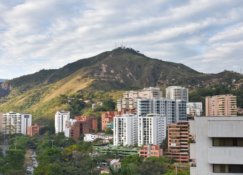 A vibrant cityscape of Cali, Colombia, with lush Andes mountains and modern buildings under a cloudy sky. Cali